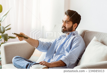 Arab Man Watching TV At Home. Young Eastern Guy With Remote Controller In Hand Resting In Living Room, Sitting On Couch And Switching Television Channels, Millennial Male Enjoying Domestic Rest 134085013