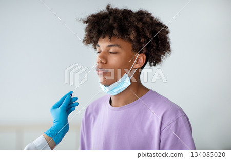 Coronavirus diagnostics. Doctor in rubber gloves making nasal coronavirus PCR test for African American teen, using sterile swab stick indoors. Black adolescent undergoing covid-19 testing procedure 134085020