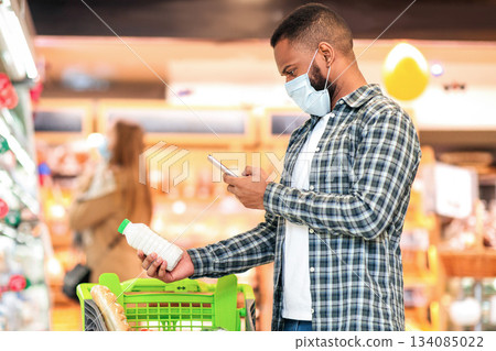 Modern Grocery Shopping. African Guy In Mask Using Phone Buying Food, Scanning Products And Using Groceries Checklist Mobile App Standing With Cart In Supermarket Store. Empty Space 134085022