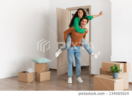 Celebrating Moving Day. Happy couple celebrating relocation to their own home, cheerful guy giving lady piggyback ride, excited young millennial family of two having fun standing in new flat 134085024