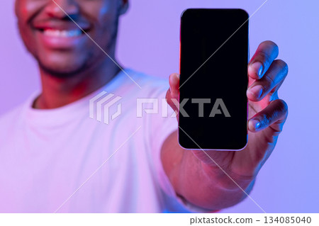 Cheerful African American Guy Showing Black Smartphone Screen With Copy Space For Your App Design, Recommending New Application, Standing In Neon Light Over Purple Studio Background, Mockup Image 134085040