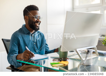 Positive black businessman writing notes in notebook while watching webinar video course, looking at computer monitor and smiling, sitting at workplace in office 134085048