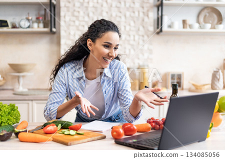 Happy latin woman talking to laptop webcamera while preparing fresh salad in kitchen interior, having video call. Food blog, social distance and recipe concept 134085056