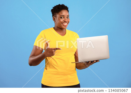 Oversized Black Woman Pointing Finger At Laptop Posing Holding Computer Advertising Website Standing In Studio Over Blue Background, Smiling To Camera. Online Service Ad Oversized Black Woman Pointing Finger At Laptop Posing Holding Computer Advertising Website Standing In Studio Over Blue Background, Smiling To Camera. Online Service Ad 134085067