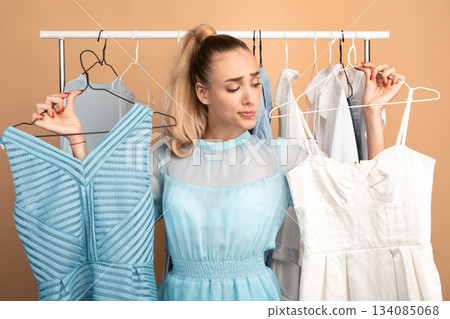 Making Choice. Portrait of young pensive woman holding two hangers with dresses and choosing trendy outfit, thinking about purchase and what to wear, standing in wardrobe, clothing rail in background 134085068
