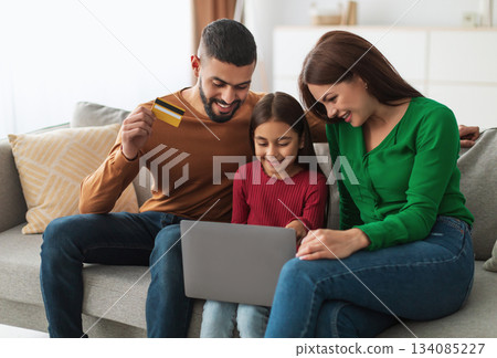 Easy Payment And Cashback Concept. Portrait of smiling Middle Eastern family doing online shopping at home using laptop pc, man holding debit credit card in hand, three people sitting on couch at home Easy Payment And Cashback Concept. Portrait of smiling Middle Eastern family doing online shopping at home using laptop pc, man holding debit credit card in hand, three people sitting on couch at home 134085227