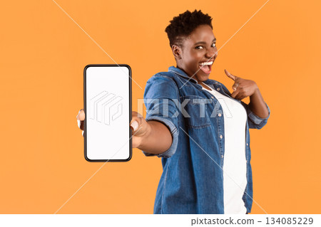 Mobile Application Ad. Excited African Woman Showing Phone Empty Screen Recommending App Standing Over Yellow Studio Background, Smiling To Camera. Cellphone Display Mockup, Check This Apps 134085229