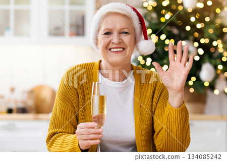 Positive mature lady in Santa hat celebrating New Year with her family or friends online, having video call, waving at camera, holding glass of champagne, decorated kitchen interior, empty space 134085242