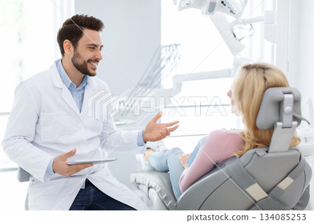 Male dentist showing female patient jaw x-ray on digital tablet, dental clinic interior. Bearded man stomatologist having conversation with lady in dental chair, holding pad, side view, copy space Male dentist showing female patient jaw x-ray on digital tablet, dental clinic interior. Bearded man stomatologist having conversation with lady in dental chair, holding pad, side view, copy space 134085253