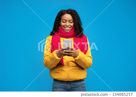 Cheerful African American Girl Using Phone Texting Online Wearing Winter Clothes Standing On Blue Background In Studio. Mobile App Concept Cheerful African American Girl Using Phone Texting Online Wearing Winter Clothes Standing On Blue Background In Studio. Mobile App Concept 134085257