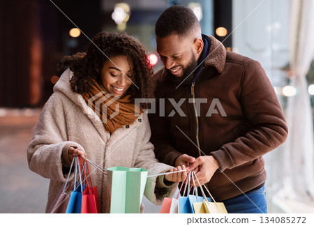 Portrait of lovely black couple holding many colorful shopping bags, looking inside, woman showing what she bought to her curious boyfriend or husband, family walking down the street in the evening Portrait of lovely black couple holding many colorful shopping bags, looking inside, woman showing what she bought to her curious boyfriend or husband, family walking down the street in the evening 134085272