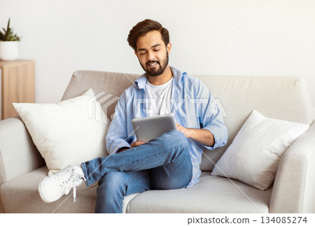 Modern Technologies For Leisure. Smiling Arab Man Using Digital Tablet At Home While Relaxing On Couch In Living Room, Eastern Millennial Guy Shopping Online Or Browsing Internet, Copy Space 134085274