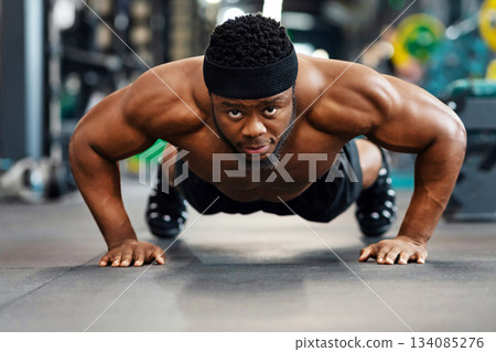 Push-ups, press-ups workout. Muscular african american shirtless guy athlete building up muscles, black man training on floor at modern gym, doing push-ups and looking at camera 134085276