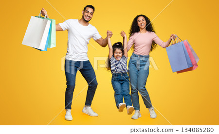 Family Shopping. Joyful Arab Parents And Little Daughter Holding Bright Shopper Bags And Having Fun Together Over Yellow Studio Background, Cheerful Mom, Dad And Child Enjoying Seasonal Sales Family Shopping. Joyful Arab Parents And Little Daughter Holding Bright Shopper Bags And Having Fun Together Over Yellow Studio Background, Cheerful Mom, Dad And Child Enjoying Seasonal Sales 134085280