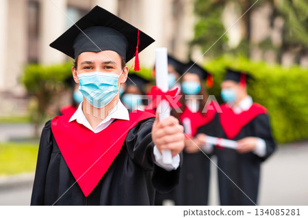 Caucasian guy student in graduation costume and protective face mask showing his diploma at camera, posing over international group of students at university campus, selective focus, copy space Caucasian guy student in graduation costume and protective face mask showing his diploma at camera, posing over international group of students at university campus, selective focus, copy space 134085281