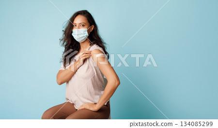 Covid-19 Vaccination. Vaccinated Lady Showing Arm After Coronavirus Antiviral Vaccine Injection Sitting Over Blue Background, Wearing Face Mask. Corona Virus Prevention. Panorama, Empty Space 134085299