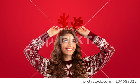 It's Christmas time. Pretty young woman in woolen sweater wearing deer horns on red studio background, panorama. Happy millennial lady having Xmas fun, celebrating New Year 134085323