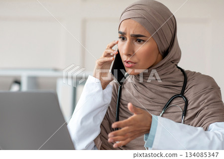 Emotional female doctor young lady in hijab looking at laptop screen, talking on phone and gesturing, having conversation with patient, giving recommendations, working at clinic Emotional female doctor young lady in hijab looking at laptop screen, talking on phone and gesturing, having conversation with patient, giving recommendations, working at clinic 134085347