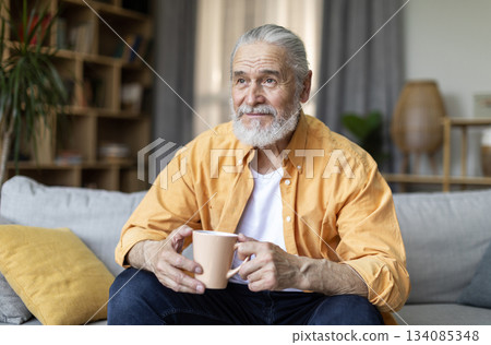 Happy grey-haired bearded senior man in stylish casual outfit drinking coffee at home, holding cup and looking at copy space for advertisement or text, living room interior Happy grey-haired bearded senior man in stylish casual outfit drinking coffee at home, holding cup and looking at copy space for advertisement or text, living room interior 134085348