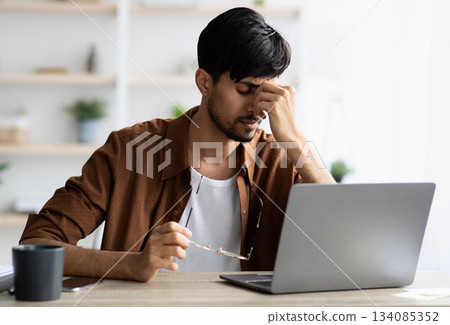 Tired indian man sitting in front of laptop at office, touching his head, suffering from burnout or financial crisis, young guy freelancer having difficulties with job, lack of clients, copy space 134085352