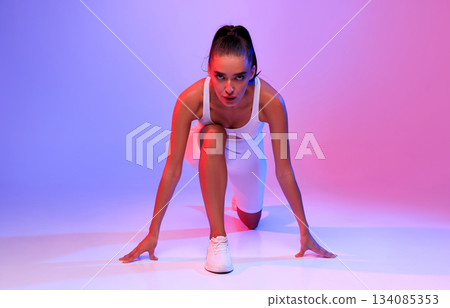 Front-View Of Motivated Female Runner Doing Crouch Start Looking At Camera Ready For Race Posing On Pink And Blue Neon Studio Background. Fit Woman Preparing For Running Workout 134085353
