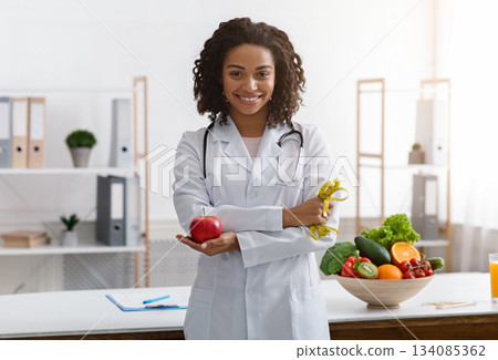 African female dietician with crossed arms holding fresh apple, posing at workplace 134085362