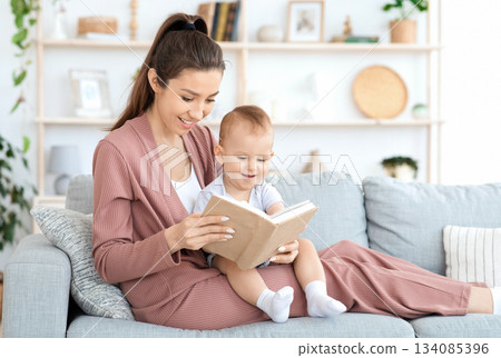Early development for babies. Millennial mom reading book to her adorable toddler baby boy at home, sitting on couch in cozy living room, free space 134085396