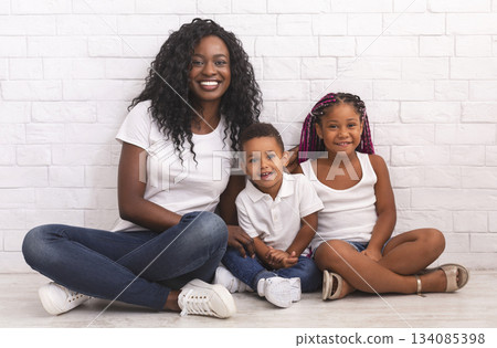 Single mother. Young african american woman with cute little kids, daughter and son, sitting on floor and posing to camera over white wall background. Single mother. Young african american woman with cute little kids, daughter and son, sitting on floor and posing to camera over white wall background. 134085398