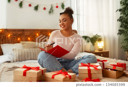 Happy african american lady writing list of presents for her family members, sitting on bed among Christmas wrapped gift boxes. Dreamy woman taking notes in notebook and smiling 134085399