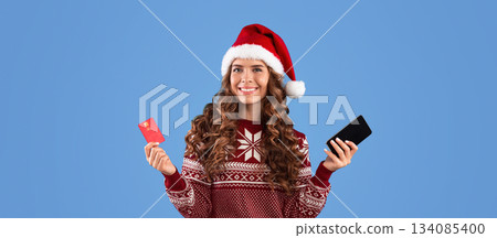 Online Christmas shopping. Lovely millennial girl in Santa hat and warm sweater holding mobile phone and credit card on blue background. Panorama Online Christmas shopping. Lovely millennial girl in Santa hat and warm sweater holding mobile phone and credit card on blue background. Panorama 134085400