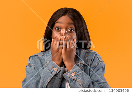 Closeup of emotional african american young woman with fair hair in jeans coat showing amazement, covering mouth with palms, orange studio background. Face expression, emotions concept 134085406