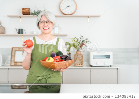 一位頭髮花白的亞洲老婦在家裡的廚房拿著蔬菜 一位頭髮花白的亞洲老婦在家裡的廚房拿著蔬菜 134085412