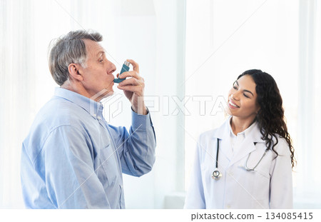 Medical Advice. Elderly man using asthma inhaler, mexican doctor watching, free space Medical Advice. Elderly man using asthma inhaler, mexican doctor watching, free space 134085415