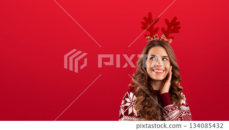 It's Christmas time. Beautiful young lady in colorful warm sweater wearing deer horns on red background. Panorama 134085432