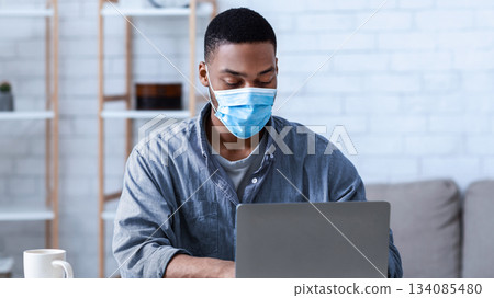 Black Man Wearing Face Mask Working On Laptop Sitting In Modern Office. Social Distancing At Work During Coronavirus Covid-19 Pandemic. Remote Work Concept. Selective Focus Black Man Wearing Face Mask Working On Laptop Sitting In Modern Office. Social Distancing At Work During Coronavirus Covid-19 Pandemic. Remote Work Concept. Selective Focus 134085480