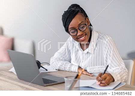 Cheerful african american business lady in formal outfit and eyeglasses attending webinar or business training, sitting at workdesk, using laptop at home, taking notes, copy space Cheerful african american business lady in formal outfit and eyeglasses attending webinar or business training, sitting at workdesk, using laptop at home, taking notes, copy space 134085482