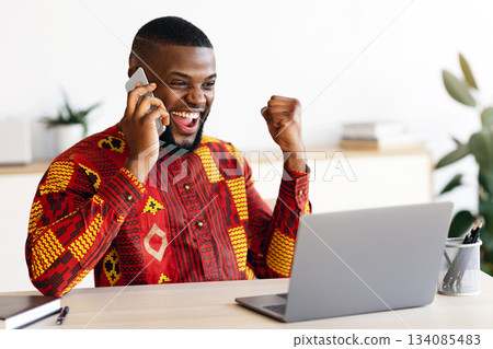 Overjoyed African Businessman In Traditional Clothes Celebrating Success At Workplace In Office, Talking On Cellphone And Looking At Laptop Screen, Emotionally Reacting To Good News, Free Space Overjoyed African Businessman In Traditional Clothes Celebrating Success At Workplace In Office, Talking On Cellphone And Looking At Laptop Screen, Emotionally Reacting To Good News, Free Space 134085483
