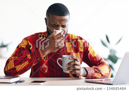 Sick Black Man In Ethnic African Shirt Sneezing At Workplace In Office, Blowing Nose To Napkin, Ill Millennial Man In Traditional Costume Suffering Seasonal Flu Or Having Coronavirus Symptoms Sick Black Man In Ethnic African Shirt Sneezing At Workplace In Office, Blowing Nose To Napkin, Ill Millennial Man In Traditional Costume Suffering Seasonal Flu Or Having Coronavirus Symptoms 134085484