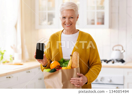 Cheerful senior woman with shopping bag full of grocery showing smartphone with blank screen, kitchen interior, copy space, mockup. Smiling old lady using mobile app for grocery delivery Cheerful senior woman with shopping bag full of grocery showing smartphone with blank screen, kitchen interior, copy space, mockup. Smiling old lady using mobile app for grocery delivery 134085494