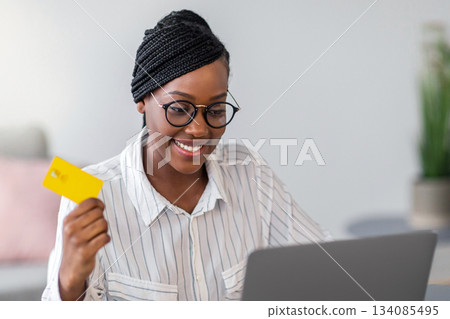 Pretty african american young woman freelancer holding credit card, using laptop, copy space. Happy black lady shopping online, ordering food, cosmetics, clothes from internet while working from home Pretty african american young woman freelancer holding credit card, using laptop, copy space. Happy black lady shopping online, ordering food, cosmetics, clothes from internet while working from home 134085495