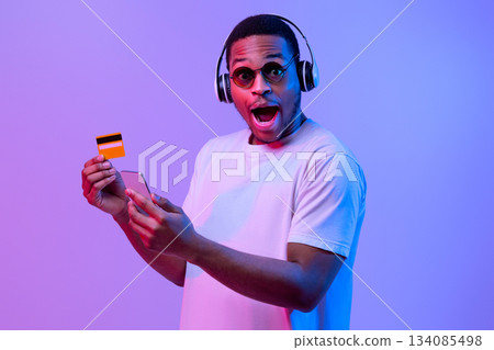 Easy Payments. Excited african american man in white t-shirt and wireless headphones holding smartphone and credit card, paying for online purchase, standing in neon light over purple background Easy Payments. Excited african american man in white t-shirt and wireless headphones holding smartphone and credit card, paying for online purchase, standing in neon light over purple background 134085498