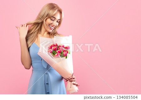 Cheerful millennial lady with bouquet of beautiful flowers on pink studio background. Pretty young blonde with bunch of tulips celebrating Woman's Day. Spring holiday concept 134085504