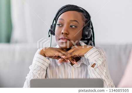 Pensive young black lady having online training at home, sitting at workdesk in front of laptop, leaning on her elbows and looking at copy space, using headphones. E-education, online class concept Pensive young black lady having online training at home, sitting at workdesk in front of laptop, leaning on her elbows and looking at copy space, using headphones. E-education, online class concept 134085505