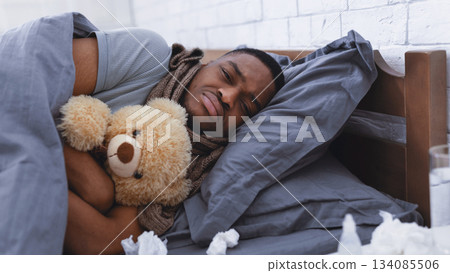 Depressed Sick African Man Hugging Teddy Bear Toy Lying Wrapped In Blanket Having Cold In Bedroom At Home. Flu And Depression, Mental Health During Coronavirus Disease Concept. Selective Focus Depressed Sick African Man Hugging Teddy Bear Toy Lying Wrapped In Blanket Having Cold In Bedroom At Home. Flu And Depression, Mental Health During Coronavirus Disease Concept. Selective Focus 134085506