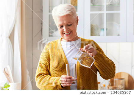 Hydration, healthy lifestyle cocnept. Smiling senior woman pouring water into glass, kitchen interior. Attractive older woman in casual outfit holding glass of fresh water, copy space Hydration, healthy lifestyle cocnept. Smiling senior woman pouring water into glass, kitchen interior. Attractive older woman in casual outfit holding glass of fresh water, copy space 134085507
