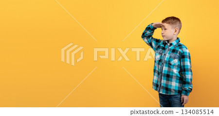 Cute ginger kid in flannel holding palm next to forehead, looking at copy space, panorama, yellow studio background. Curious red-haired boy watching exciting advertisement for kids 134085514