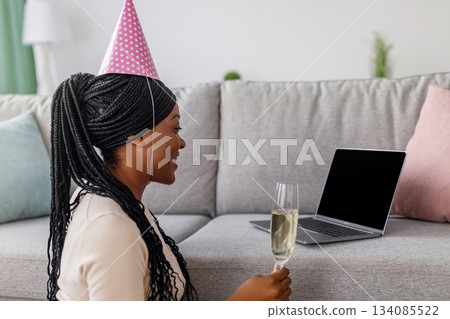 Young african american lady having online birthday party from home during coronavirus pandemic, wearing party hat, holding glass of champagne and looking at empty laptop screen, copy space Young african american lady having online birthday party from home during coronavirus pandemic, wearing party hat, holding glass of champagne and looking at empty laptop screen, copy space 134085522