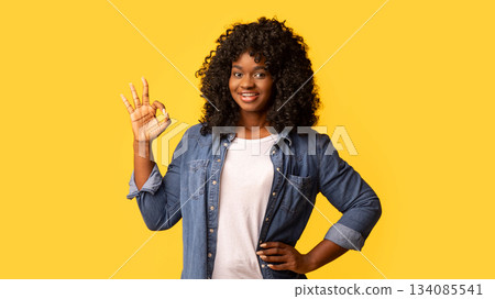Happy beautiful black woman with curly hair standing on yellow studio background, showing okay gesture, copy space. Happy and carefree african american young lady in casual jeans shirt enjoying life 134085541