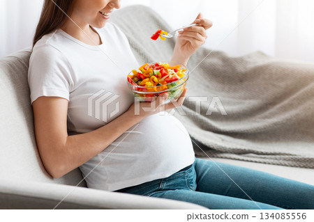 Vitamin Food. Happy Young Pregnant Woman Eating Vegetable Salad While Sitting On Couch At Home, Relaxing In Living Room And Enjoying Healthy Organic Nutrition During Pregnancy, Side View, Copy Space Vitamin Food. Happy Young Pregnant Woman Eating Vegetable Salad While Sitting On Couch At Home, Relaxing In Living Room And Enjoying Healthy Organic Nutrition During Pregnancy, Side View, Copy Space 134085556