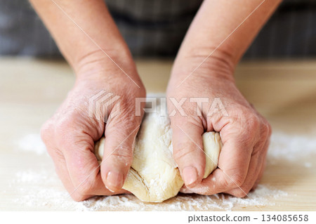 Culinary, cooking, baking concept. Female hands kneading dough, unrecognizable woman grandmother in apron cooking in kitchen, making pastry, cookies, cake or pie, pizza making, cropped Culinary, cooking, baking concept. Female hands kneading dough, unrecognizable woman grandmother in apron cooking in kitchen, making pastry, cookies, cake or pie, pizza making, cropped 134085658